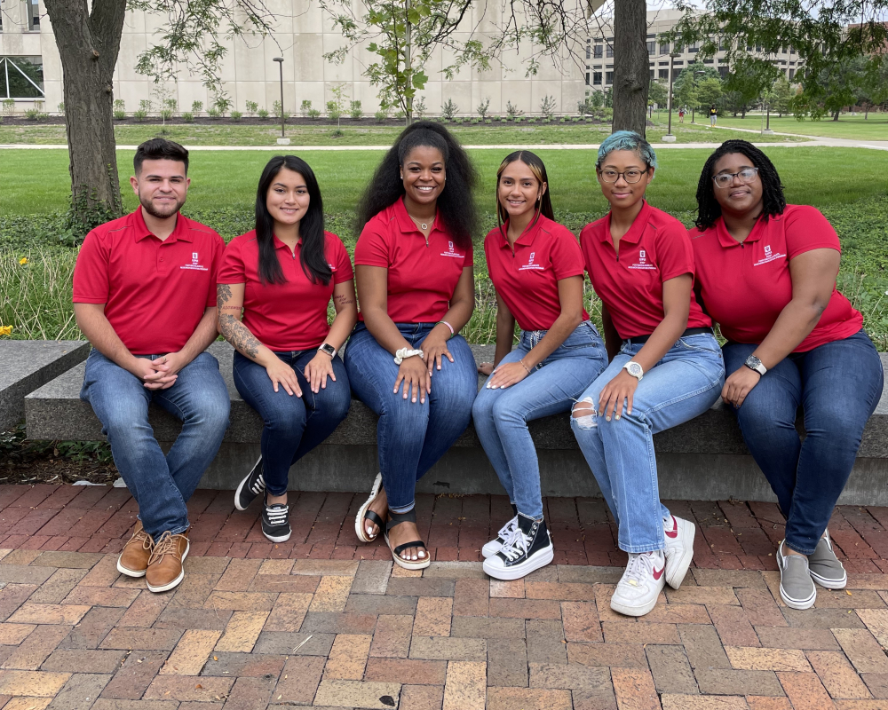 The 2022 IPREP fellows, from left, are Andres Prieto Trujillo, Selena Martinez, Ceouna Hegwood, Lorimar Robledo Gonzalez, Rieanna McPhie and Eriel Wise. They are all wearing matching red polos and blue jeans sitting on a bench in a greenspace. 
